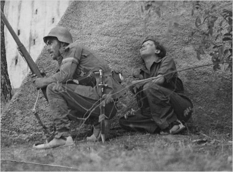 Gerda Taro dietro un soldato 
		   durante una battaglia (foto di Robert Capa)